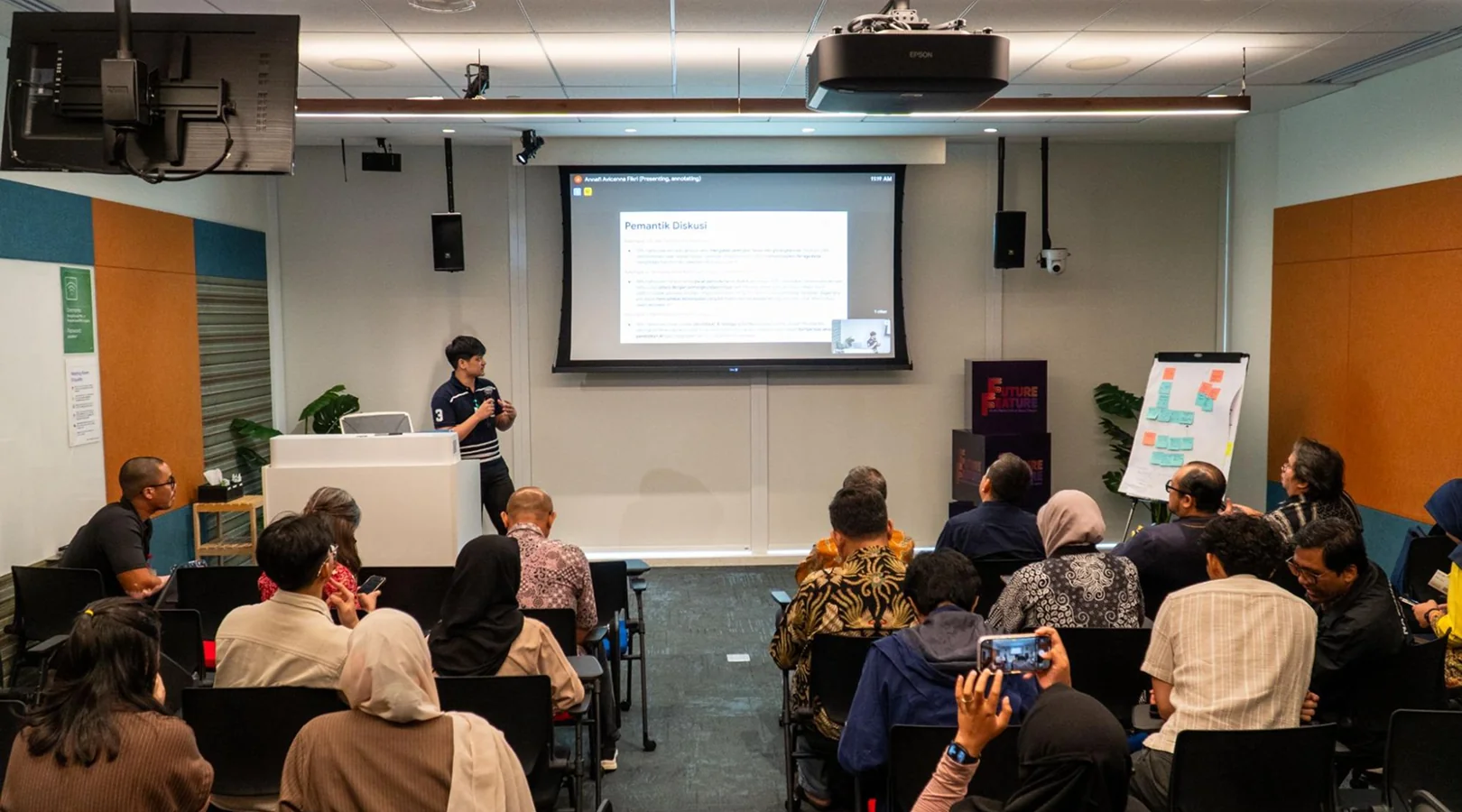 Muttaqin standing at a lectern presenting to a seated audience, with a slide titled “Pemantik Diskusi” on screen